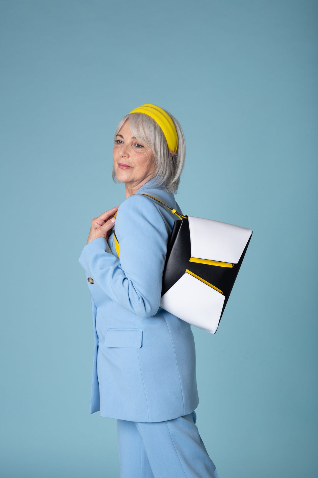 Senior woman in stylish light blue suit and yellow headband holding a modern white and black handbag against a blue background