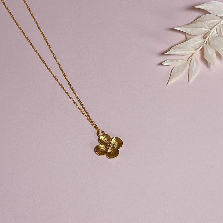 Gold flower pendant necklace on a light pink surface with white leaves