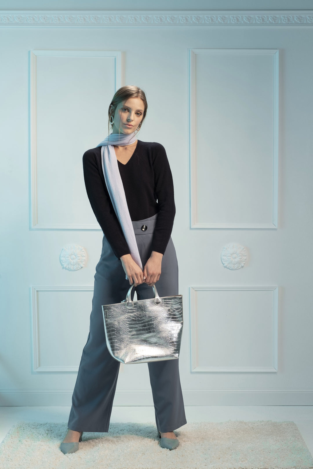 Woman in chic outfit holding silver handbag against decorative white wall